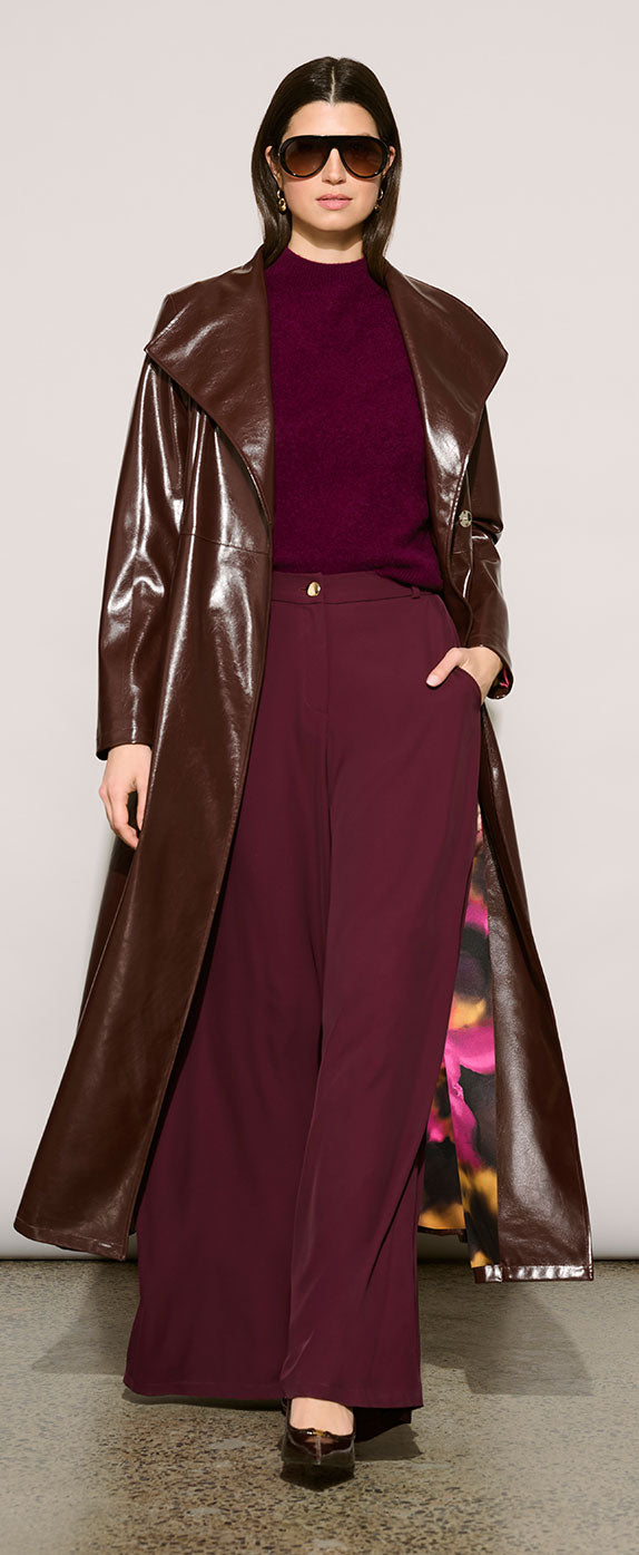 A model wearing Joseph Ribkoff Edit 57 long brown faux-leather coat over a deep burgundy knit top and matching wide-leg trousers