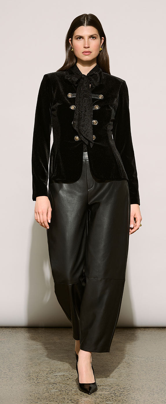 A model wearing Joseph Ribkoff Edit 57 black velvet cropped jacket with gold buttons and a black satin bow collar, paired with black leather pants